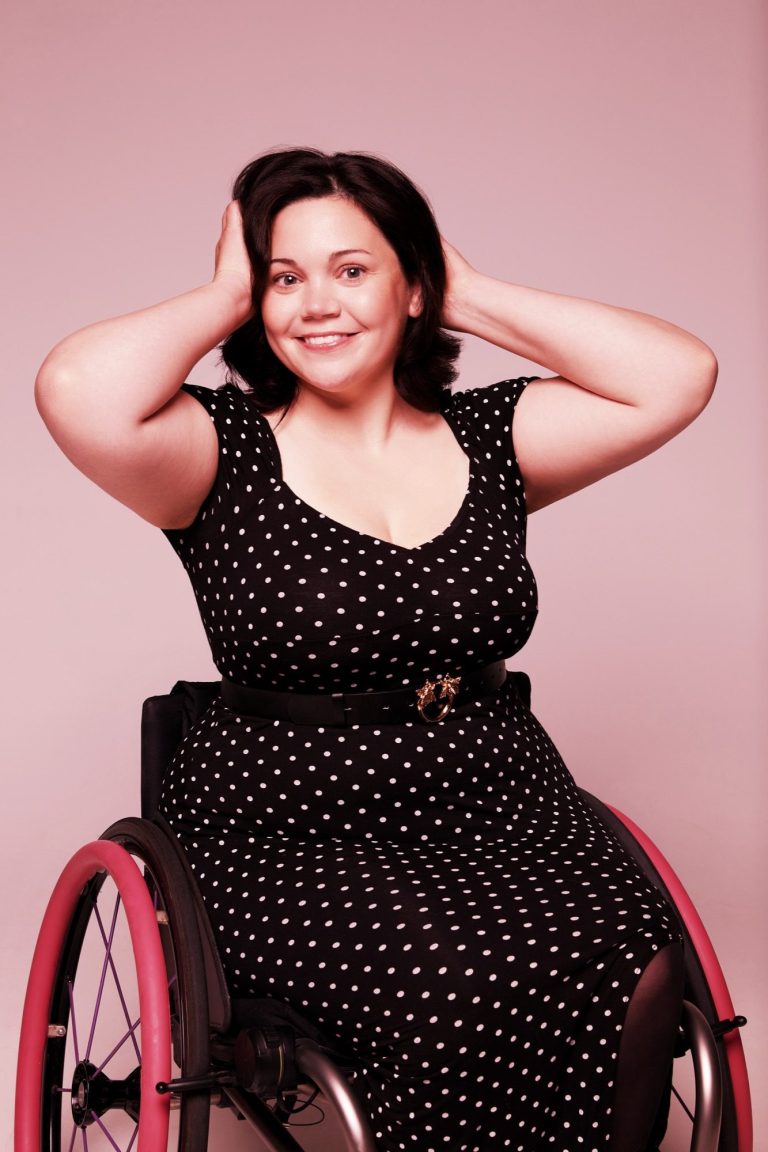 Smiling woman in a wheelchair wears a polka dot dress, hands on her head against a pink background.
