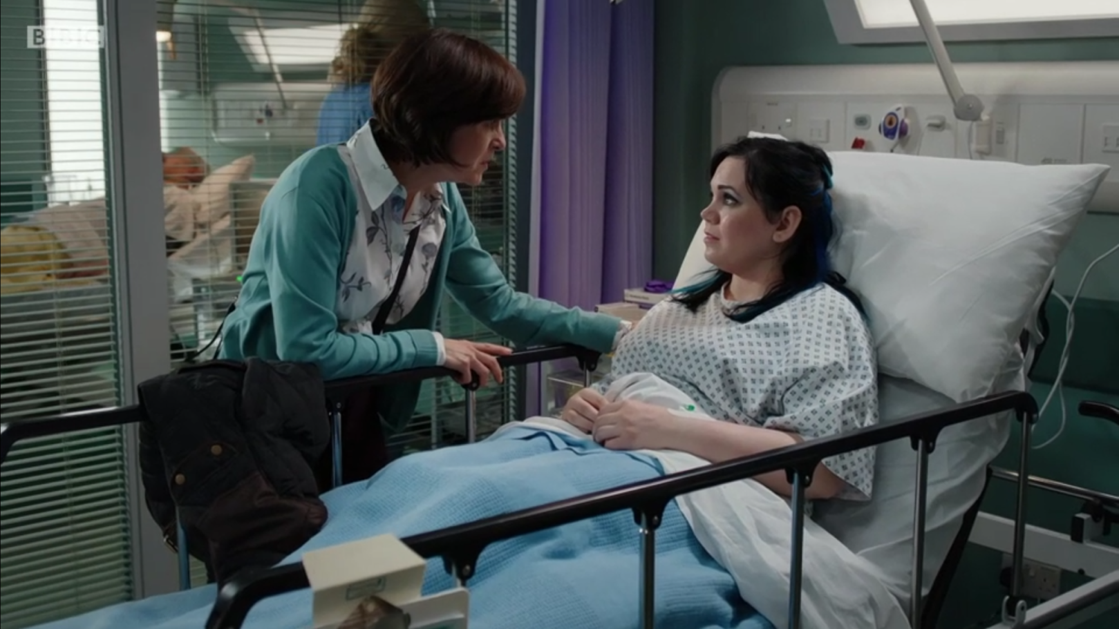 Hannah is on set in a hospital bed. She has a hospital gown and looking sassily at her screen mum who looks on in a concerned way